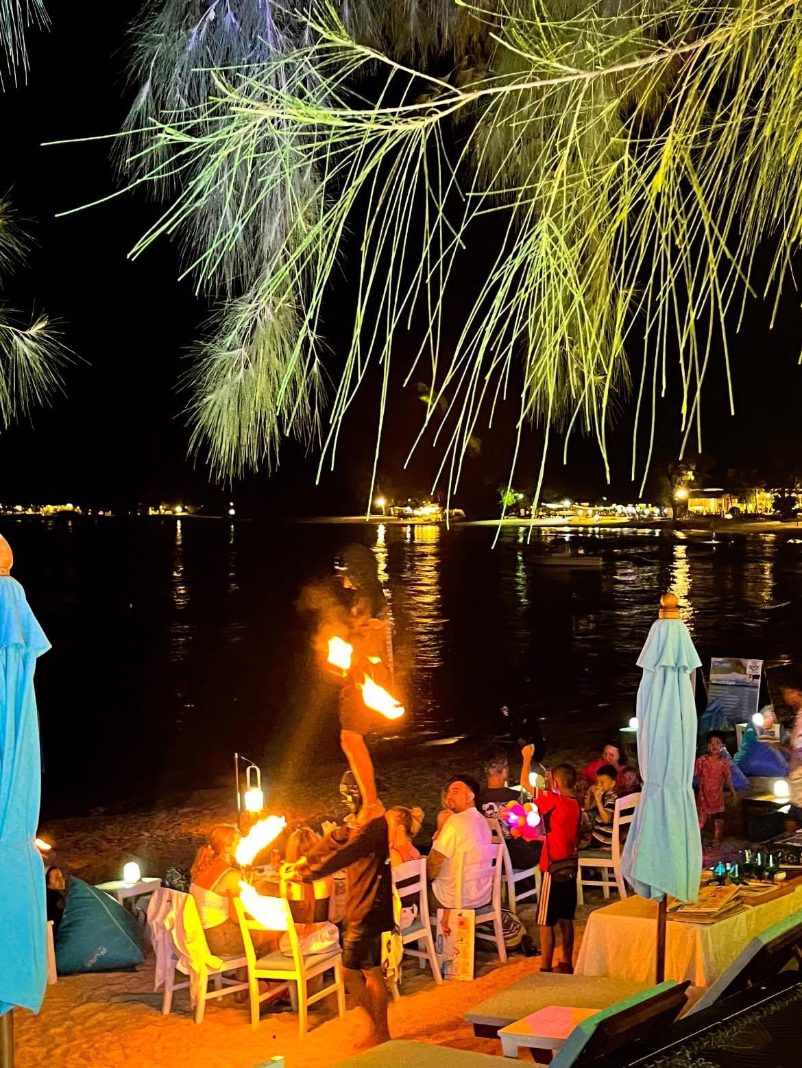 Fire performer between the tables — fireworks lighting up the bay behind
