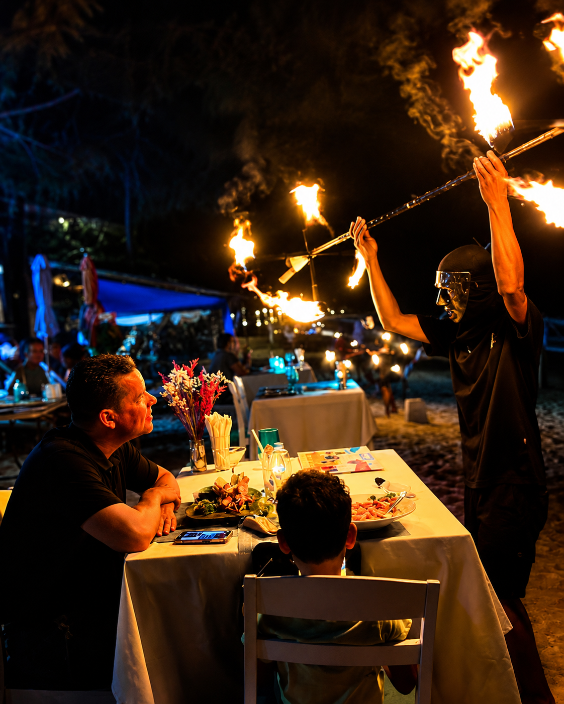 Fire performer holding flaming staffs beside guests dining on the beach