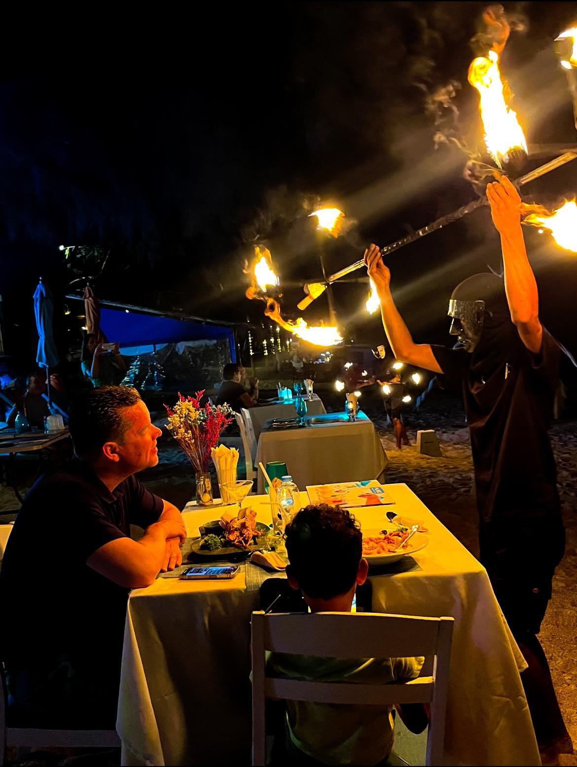 Fire show performer at a dining table — guests watching from their beachfront seats at The Blue Spice